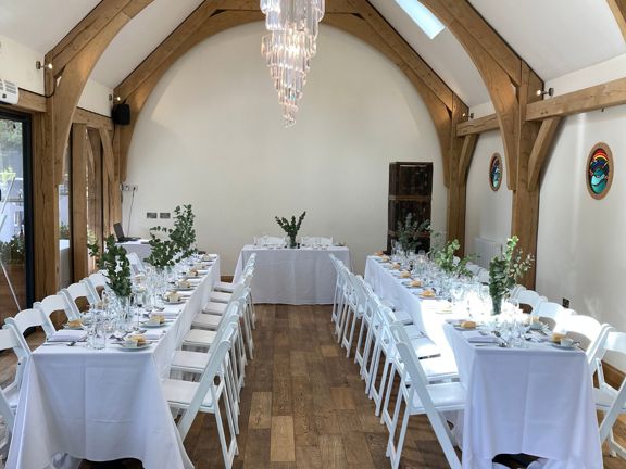 Barn with wooden beams, wooden flooring, white walls and glass chandelier. White chairs with tables set up for a meal in long rows.
