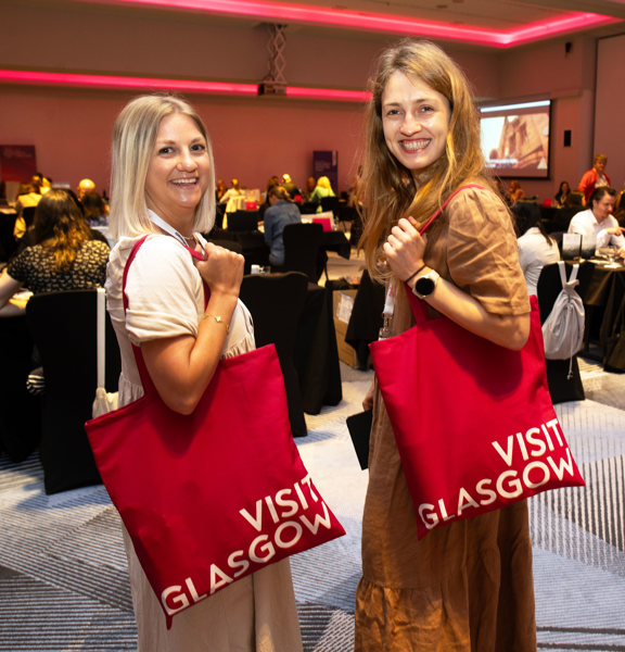 Two people looking over their shoulder with a Visit Glasgow tote back on it. 