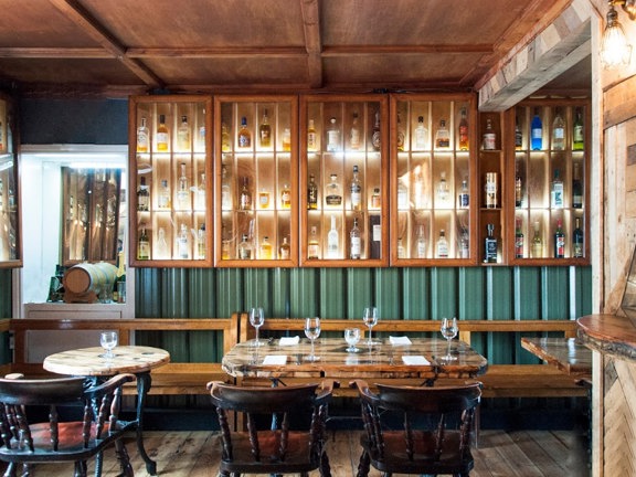 An interior view of a dining room at The Finnieston restaurant. A bright room with refinished wooden floorboards and walls decorated with reclaimed wood panelling and green corrugated metal. Light varnished pigeonholes line the far and left walls, they are lit and glass fronted – displaying bottles of whisky. A long varnished wooden bench, that lines the base of both walls, and dark wood, round-back chairs are arranged around wooden tables, with cast iron legs, set for 2 – 4 people.