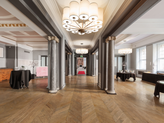 Interior view of grand hotel foyer. Herringbone wooden floors, marble pillars and occasional buffet tables with black table cloths.