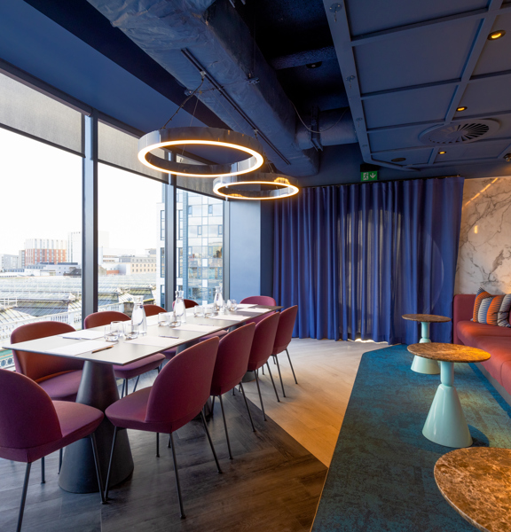Meeting room in a hotel, a red/brown banquet stretching the length of the right hand side of the room facing a long white table surrounded by 8 red chairs looking out floor to ceiling windows to the left of the room. 