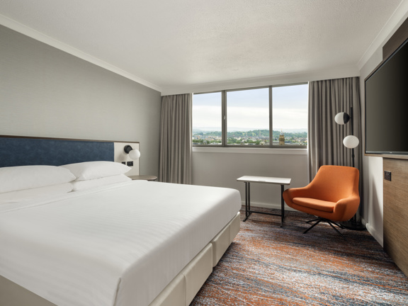 Hotel bedroom with one king-size bed dressed with crisp white linen and 4 fluffy white pillows. An orange accent chairs sits in the corner under a large window looking out across the city. 