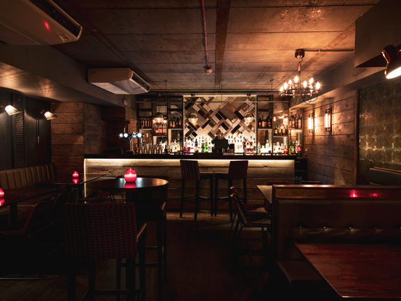 The darkly decorated bar at blue dog. High tables and booths in dark woods and leather can be made out. A bar with rustic wooden planks decorating its front is lit at the end of the room. The mirrored shelves behind are well stocked. A chandelier, wall sconces and candles on the table offer low light.