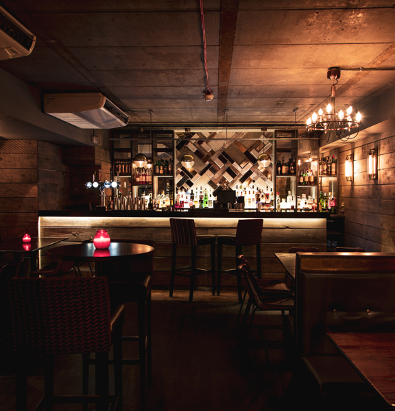 The darkly decorated bar at blue dog. High tables and booths in dark woods and leather can be made out. A bar with rustic wooden planks decorating its front is lit at the end of the room. The mirrored shelves behind are well stocked. A chandelier, wall sconces and candles on the table offer low light.