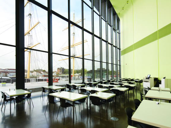 An interior view of the the Riverside Museum cafe space, a huge 2-storey glass wall with dark frames dominates the space. Outside a paved area with railings and 2 large, yellow ship masts are visible. Inside small white tables are arranged in small groups with black, wire legged chairs. The interior wall is a pale green and has spotlight mounted to it. A till point can  be seen in the bottom right corner.