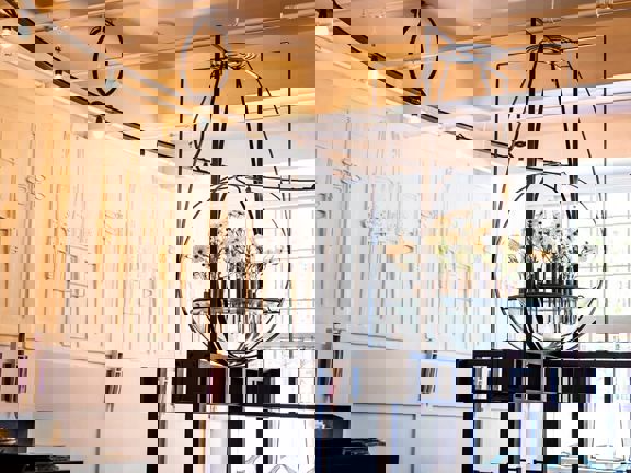 An ornate mental structure hangs from the ceiling. At the entre is a class vase filled with white flowers. The walls are ornately decorated with plasterwork in the Charles Rennie Mackintosh style