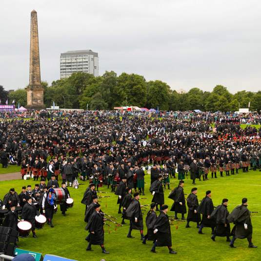Hundreds of pipers in dark kilts and clothes march across Glasgow Green with merchandise and food stands in the background