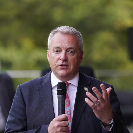 Man in a dark suit and patterned tie speaking into a handheld microphone outdoors, gesturing with one hand, with a blurred green background.