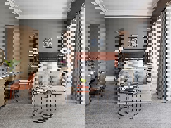 An interior shot of a Blythswood Square hotel room, a room with soft pale-grey carpet, grey walls and white plaster work, 2 windows surrounded by white wooden panelling are evident on the left wall. A large bed with a brown leather headboard made with crisp white sheets is against the far wall. a pale grey loveseat is at its foot. 2 small, round glass nesting coffee tables, with black metal bases, sit in the foreground. Against the left wall are a modern, ashen-wood wardrobe and desk & a leather chair.