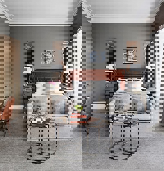 An interior shot of a Blythswood Square hotel room, a room with soft pale-grey carpet, grey walls and white plaster work, 2 windows surrounded by white wooden panelling are evident on the left wall. A large bed with a brown leather headboard made with crisp white sheets is against the far wall. a pale grey loveseat is at its foot. 2 small, round glass nesting coffee tables, with black metal bases, sit in the foreground. Against the left wall are a modern, ashen-wood wardrobe and desk & a leather chair.