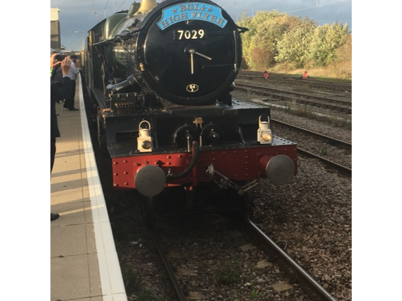 The front of a traditional steam train, bearing a sky-blue metal name plate reading "BGL High Flyer". The locomotive's panels are dark green with black, brass and red details. The train is stationary at an outdoor platform. Trees, shrubs and grass can be seen on the far right side, beyond several sets of tracks. The white edge of a platform is visible on the left-hand edge of the image, people can just be seen getting on the train and taking photos of it. Overhead wires dissect the blue, cloudy sky.