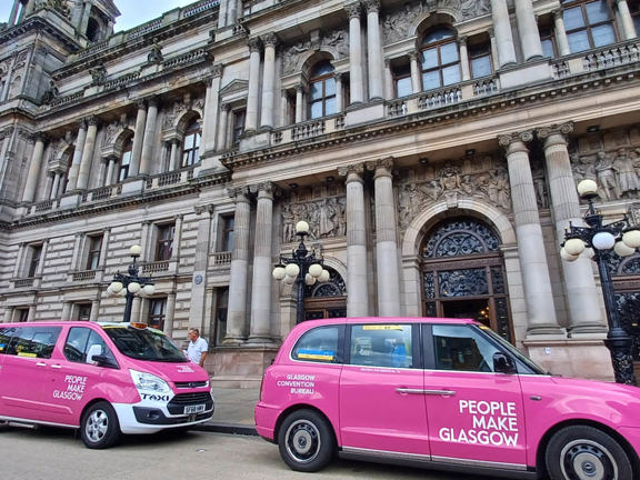 3 pink taxis with People Make Glasgow branding lined up in front of the Victorian-style Glasgow City chambers