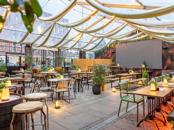 Covered terrace in Wunderbar West End. Wooden tables and chairs, ambient lighting and festoon lights hanging from the ceiling.