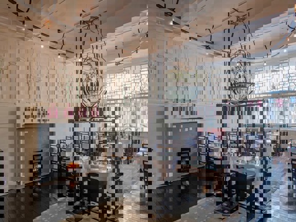 Traditional tearooms ornately plastered walls painted in cream with accents of mauve surround tables that are laid with crip white linen and delectate blue and white cups and saucers. Above an ornate metal and glass structure hangs from the ceiling  and is filled with white flowers.