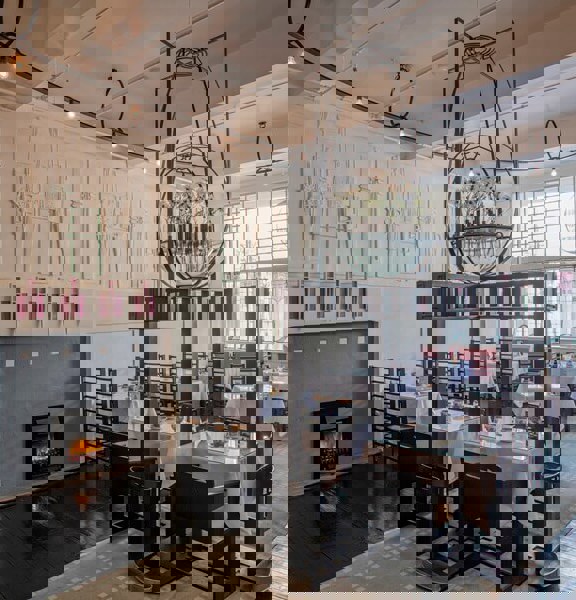 Traditional tearooms ornately plastered walls painted in cream with accents of mauve surround tables that are laid with crip white linen and delectate blue and white cups and saucers. Above an ornate metal and glass structure hangs from the ceiling  and is filled with white flowers.