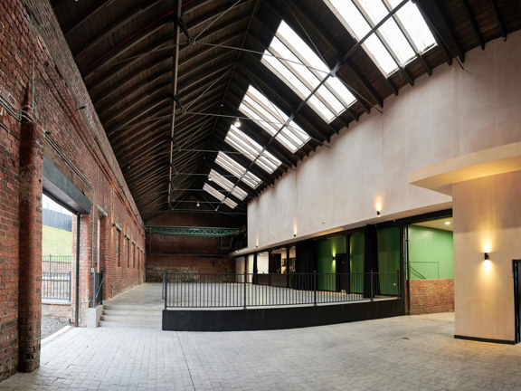 An interior image of The Engine Works main space, the Engine Room. The image shows an ex-industrial space with a mix of red brick and gypsum walls and a vaulted ceiling with skylights. The main space is over 2 levels, the lower space is paved with bricks, the 4 stairs and the upper level appear to be concrete. A black railing separates the levels. Corridors and rooms can be seen to the right, off of the main space, while an open doorway to the right reveals a sliver of a gravelled and grassy outside area.