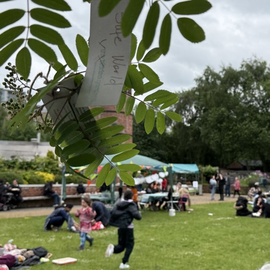The image focusses on some leaves hanging from a tree with a handwritten message tied on. In the background, groups of people are having a picnic in the gardens