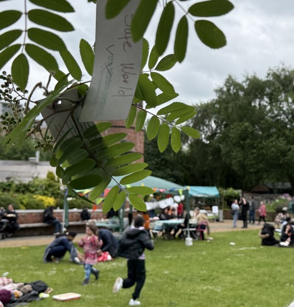 The image focusses on some leaves hanging from a tree with a handwritten message tied on. In the background, groups of people are having a picnic in the gardens
