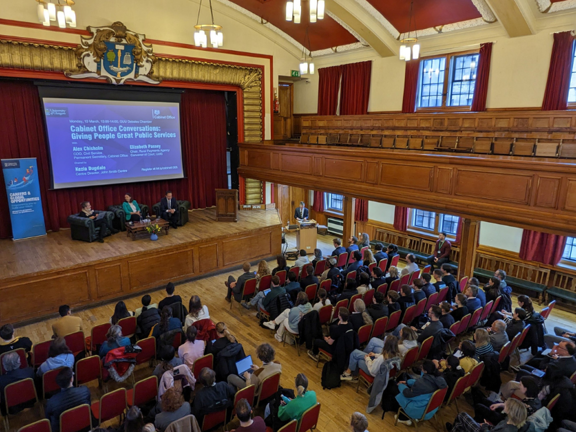 Large theatre style room with balcony and stage. On the stage there is a large digital screen with three people sat on armchairs below the screen on stage. Off stage the audience sits in rows of red chairs. 