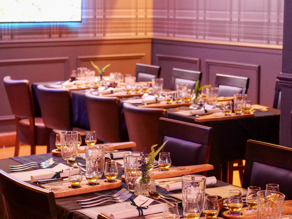 An interior view from Mharsanta shows a room decorated with dark panelling and grey, tartan wallpaper. Dark wood and leather chairs can be seen around 2 rectangular dining tables. Small glasses of whisky, cut flowers and silverware decorate the dark tablecloths. On the far wall the bottom corner of a bright television screen or projection can be interpreted.