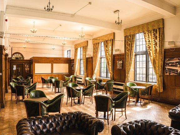 Large traditional room with parquet flooring, wooden panelling and five large windows along one length. There are green leather armchairs around wooden tables throughout the room and three brown leather chesterfield style sofas in the foreground. 