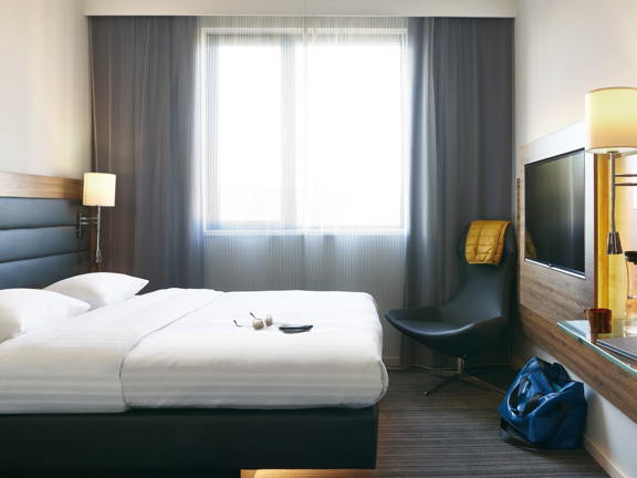An interior view of a Moxy Merchant City bedroom shows a minimal, modern hotel room with grey, striped carpet. The double bed is against the left wall, it's made with crisp white sheets and has a padded, dark-grey headboard, the beds base is a pedestal and light emits from under the bed. The far wall is taken up with a large window, with net and opaque curtains. A large TV is visible on the right wall and a modern, leather armchair is in the corner beyond. Bedside lamps and a plug socket are visible.