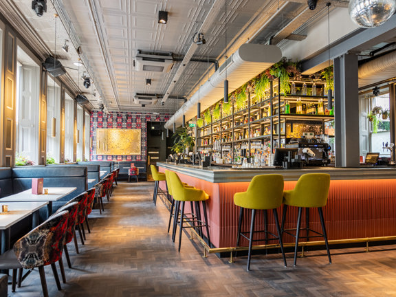 Interior of Kong bar in Glasgow featuring a industrial, modern space with banquet seating and yellow bar stools in front of a peach bar. Daylight streams in through the large windows, hanging greenery, and a well-stocked bar.  