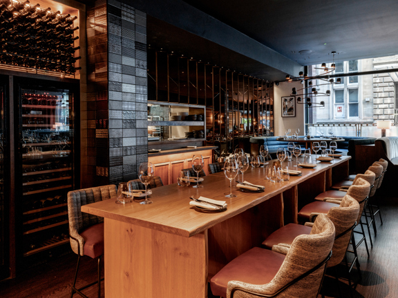 A view of Gaucho's main dining room. A darkly painted room with wood effect floors and a plate glass window on the far right wall, looking out on a Glasgow street and sandstone buildings. A tall, wooden table surrounded by leather upholstered bar stools is set for 12 diners. A fully stocked wine fridge, a hatch to the kitchen and booths can be seen lining the walls behind. A stylish, twisting light feature hangs form the ceiling in the window.