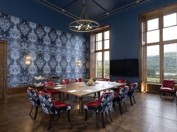 Large room with floor to ceiling windows, blue accent wallpaper. In the middle of the room is a wooden dining table surrounded by red leather chairs and with blue velvet backs.