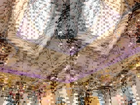 An interior view of Corinthian Club looking up at a large glass dome. Opulent, decorative plaster work is visible around the dome and across the entire ceiling in white and gold. The room is very bright with daylight streaming through the dome as well as the windows that cover both visible walls. There are also chandeliers and more modern light fittings visible. Large vases of foliage and pink flowers are also visible.