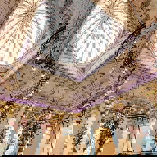 An interior view of Corinthian Club looking up at a large glass dome. Opulent, decorative plaster work is visible around the dome and across the entire ceiling in white and gold. The room is very bright with daylight streaming through the dome as well as the windows that cover both visible walls. There are also chandeliers and more modern light fittings visible. Large vases of foliage and pink flowers are also visible.