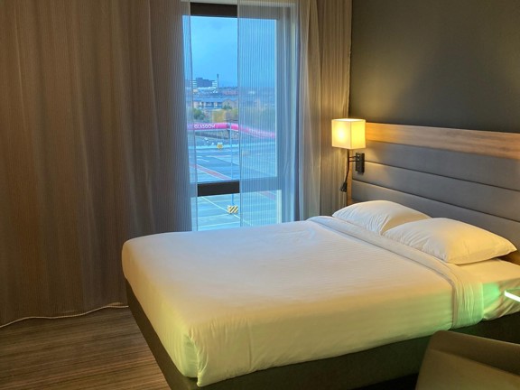 An interior view of a Moxy SEC bedroom shows a minimal hotel room with grey, striped carpet. The double bed is against the right wall, it's made with crisp white sheets and has a padded, pale-grey headboard, the beds base is a pedestal and light emits from under the bed. The far wall is taken up with a large window, with net and opaque curtains pulled back to reveal a view of the SEC campus covered walkway. A bed side lamp and a pale-grey, leather armchair are also visible on either side of the bed.