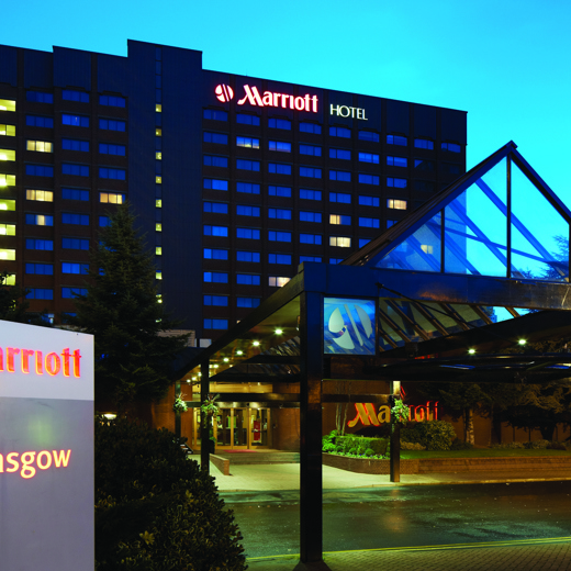 An exterior view of the Glasgow Marriott hotel at night shows a multi-storey square building. Leading to the front door is a long glass covered walkway. In the foreground there is a lit sign reading "Marriott Glasgow" and at the top of the building lit letters read, "Marriott Hotel". Shrubs and landscaping are evident on either side of the walkway.