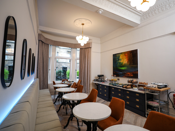 Bright and light hotel breakfast room. Long banquet runs along one wall, white circular tables and burnt orange velvet chairs sit opposite. Black unit with gold handles is mounted with a white counter which is laden with as array of breakfast pastries, juices and coffee. 