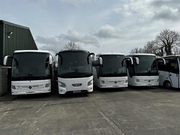 The image shows the fronts of 5 large white coaches parked side by side. On the left of them, the edge of a large grey corrugated building, which could be interpreted as a garage is visible, just on the edge of the frame. Bare trees can be seen beyond the coaches against a pale, cloudy sky.