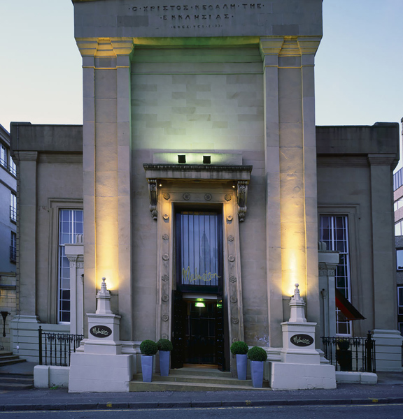 An exterior image of the Malmaison Glasgow shows a sandstone building with a strikingly, tall Egyptian-influenced façade. An enormous, carved archway houses the entrance, a black double doorway with a large window above it. Gold lettering above the door in the signature branding reads, "Malmaison." The façade is uplit with white and green lights, exaggerating its height and showcasing the carved pillars and a Greek inscription at the very top. Black railings and potted shrubs decorate the steps.