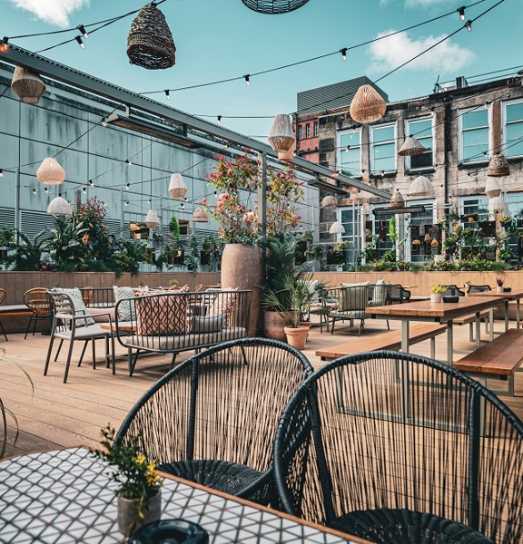 Open air terrace in Kong Glasgow. Mix of wooden and tiled tables and chairs, hanging and planted greenery and natural fabric pendants hang from above. 