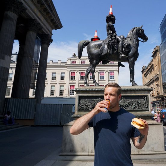 Tour Guide Steven Halcrow standing in front of the Duke Of Wellington statue in Glasgow drinking Irn Bru and holding a roll and fritter. 