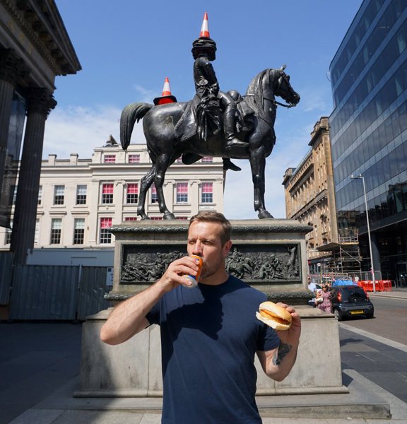 Tour Guide Steven Halcrow standing in front of the Duke Of Wellington statue in Glasgow drinking Irn Bru and holding a roll and fritter. 