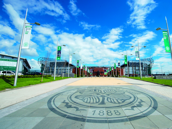 An external view of Celtic Football Club shows a paved walkway, lined with lampposts bearing green and white banners and a landscaped lawn. In the foreground the paved walkway is decorated with the Celtic Football Club crest, boasting a 4-leafed clover and the date 1888. At the end of the walkway a large, modern, red brick stadium is visible against a blue, cloudy sky.