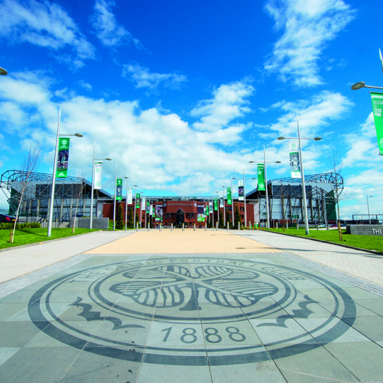An external view of Celtic Football Club shows a paved walkway, lined with lampposts bearing green and white banners and a landscaped lawn. In the foreground the paved walkway is decorated with the Celtic Football Club crest, boasting a 4-leafed clover and the date 1888. At the end of the walkway a large, modern, red brick stadium is visible against a blue, cloudy sky.