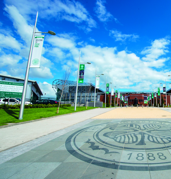 An external view of Celtic Football Club shows a paved walkway, lined with lampposts bearing green and white banners and a landscaped lawn. In the foreground the paved walkway is decorated with the Celtic Football Club crest, boasting a 4-leafed clover and the date 1888. At the end of the walkway a large, modern, red brick stadium is visible against a blue, cloudy sky.