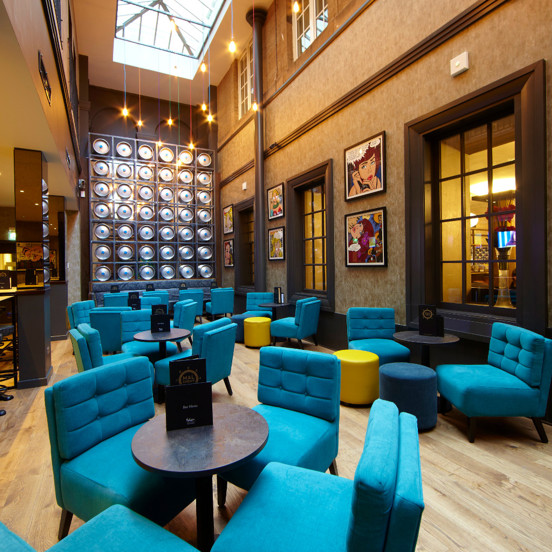 An interior view of the Malmaison Bar, shows a narrow, double-height room with a sky-light roof. A metallic art work of discs takes up most of the far wall. Low, velvet chairs and footstools in turquoise, teal and yellow surround small dark, bar tables. A lower ceilinged seating area can be seen through 2 mirrored pillars to the left. Framed prints and dark-wood internal windows and a picture rail adorn the right wall.