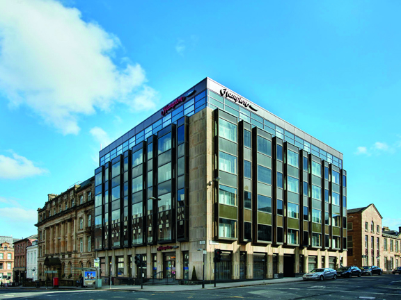 An exterior view of the Hampton by Hilton on a sunny day shows the corner of a square concrete multi-storey building with large windows on each floor and metallic details. The Hampton branding is shown at the top of each side of the building, in red. Roads can be seen stretching passed both sides of the building, there are parked cars and sandstone buildings in each direction and a bus stop and a latter box on the left. The sky is blue and bright.