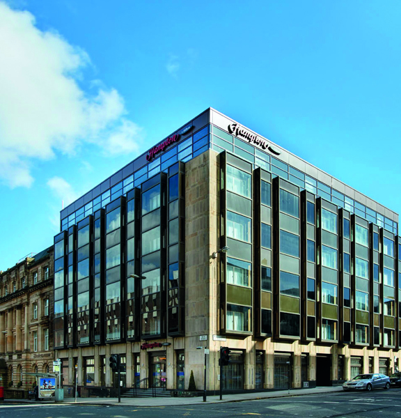 An exterior view of the Hampton by Hilton on a sunny day shows the corner of a square concrete multi-storey building with large windows on each floor and metallic details. The Hampton branding is shown at the top of each side of the building, in red. Roads can be seen stretching passed both sides of the building, there are parked cars and sandstone buildings in each direction and a bus stop and a latter box on the left. The sky is blue and bright.