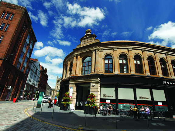 A wide lens image of the exterior of Merchant Square. A curved 2-storey Victorian building on a paved street corner with a wide pavement. The entrance is a large, square, open entrance-way on the corner; large planters sit on either side. A large arched window with carved pillars and a decorative finial sits above it. The first floor is lined with arched windows while the ground floor houses a shop front, people sit at bistro tables outside. Multiple large brick buildings can be seen over the road.