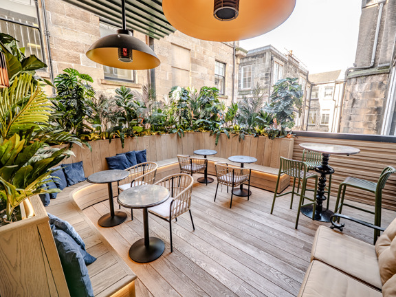 Outside terrace with occasional tables and chairs, wooden flooring and greenery. 