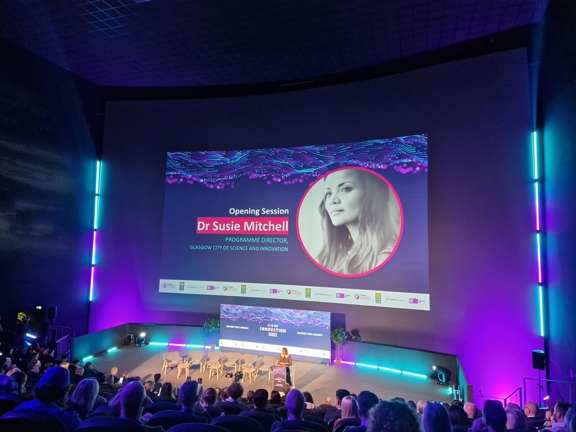 Giant Imax screen displaying conference speakers name and picture. Speaker on stage in front of screen facing audience seated in rows. 