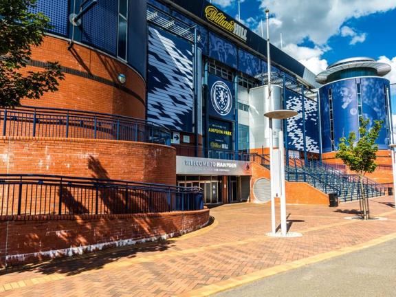 An exterior view of Hampden Stadiums main entrance. A large brick and blue metal clad building, with round towers at the two visible corners. The facade is decorated with sponsors, the Scottish FC badge and lettering in white or yellow reading," Hampden Park."  The area outside of the main entrance is paved with brick and curves down to 3 or more doors. Adjacent steps lead up to a further entrance, furthermore, ramps curve out of sight around the towers' bases. Trees and modern streetlights line the path.