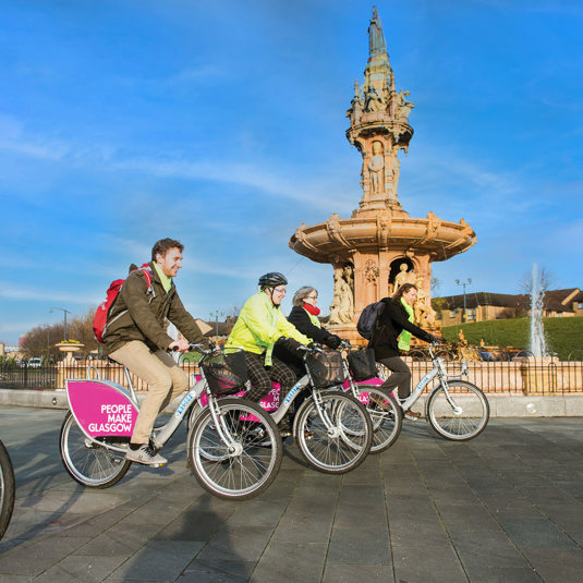Five cyclists on People Make Glasgow branded bikes in front of the large terracotta Doulton Fountain